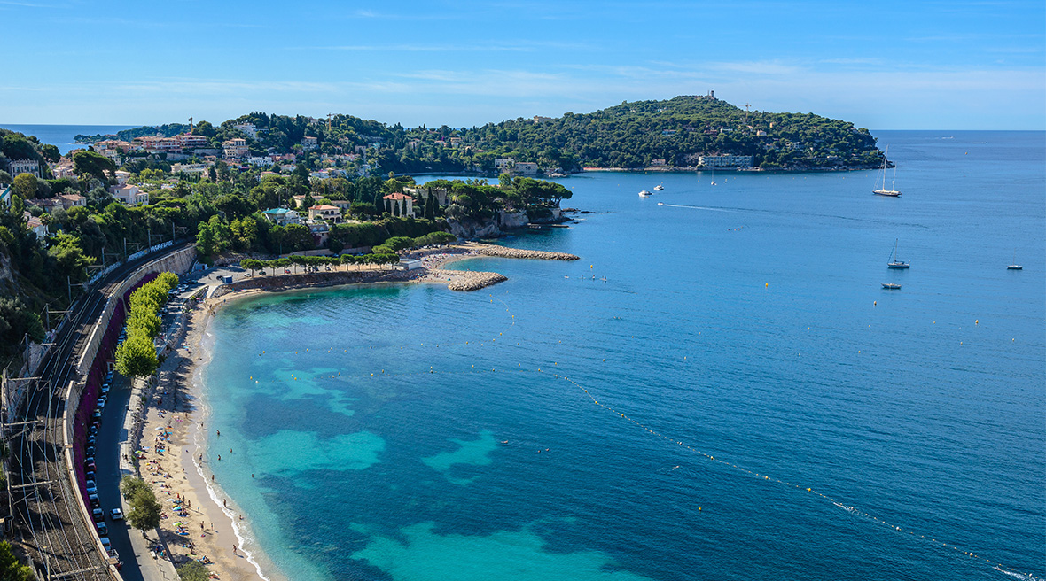 A view of the French Riviera from a road up in the cliffs that surround a bay of clear blue water and sandy beaches
